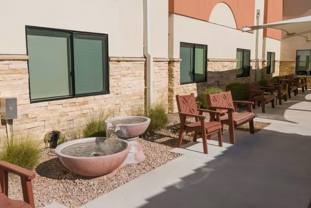 Outdoor patio area with multiple wooden benches and chairs arranged along the side of a building with stone and stucco walls. There are two round water fountains surrounded by small rocks and some plants near the building wall.