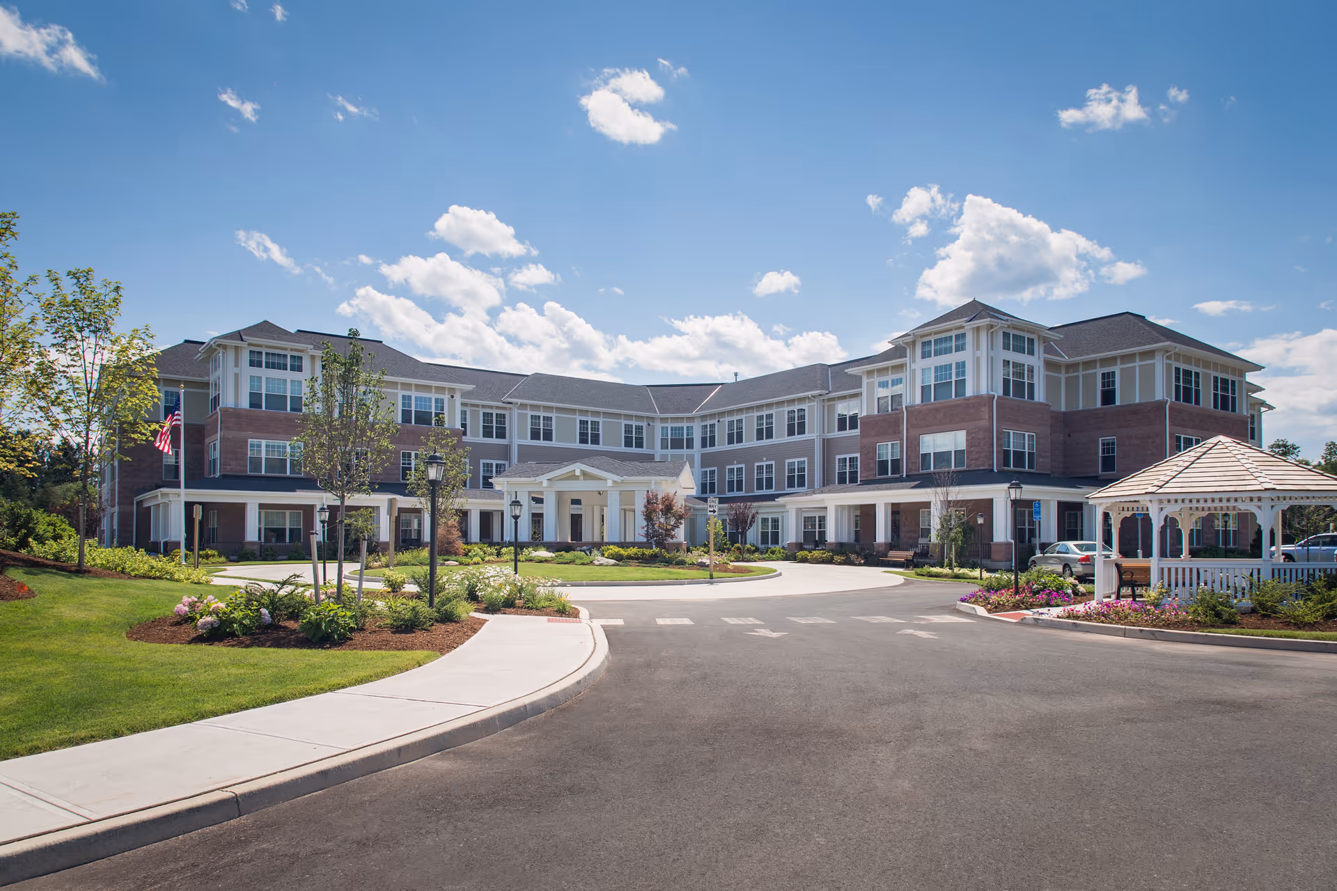 Exterior view of a large, three-story senior living facility building with brick and white siding, surrounded by landscaped gardens, a gazebo, and a driveway under a blue sky with scattered clouds.