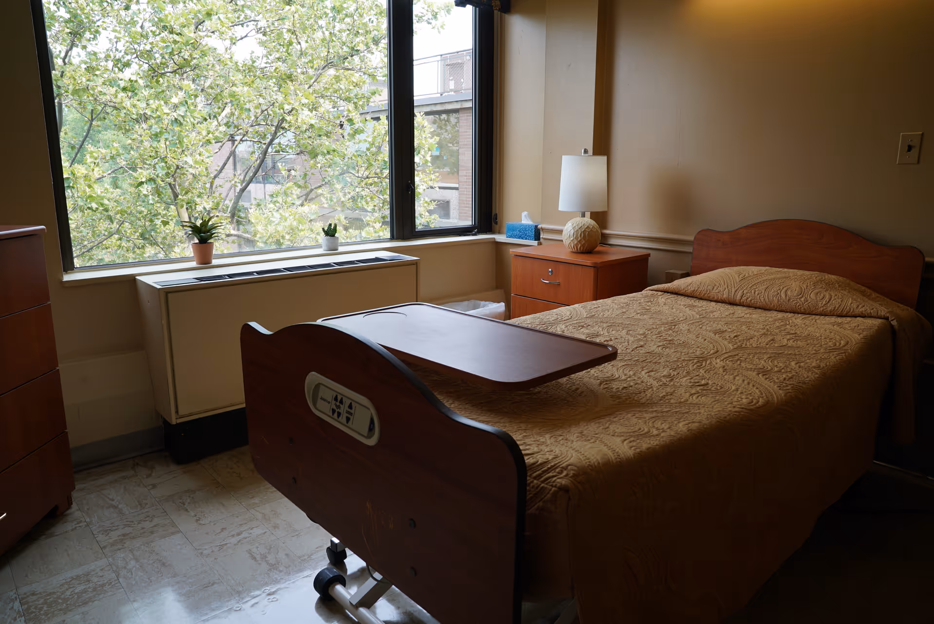 Single-occupancy bedroom with a hospital-style bed, overbed table, nightstand and a large window overlooking trees.