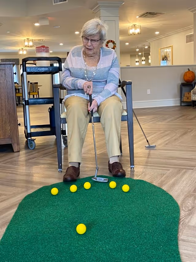 An elderly woman sitting on a chair indoors playing mini golf on a small green putting mat with several yellow golf balls. She is holding a putter and appears focused on the game. The room has wooden flooring, a cart with items on it, and some furniture including a table with a pumpkin on it in the background.