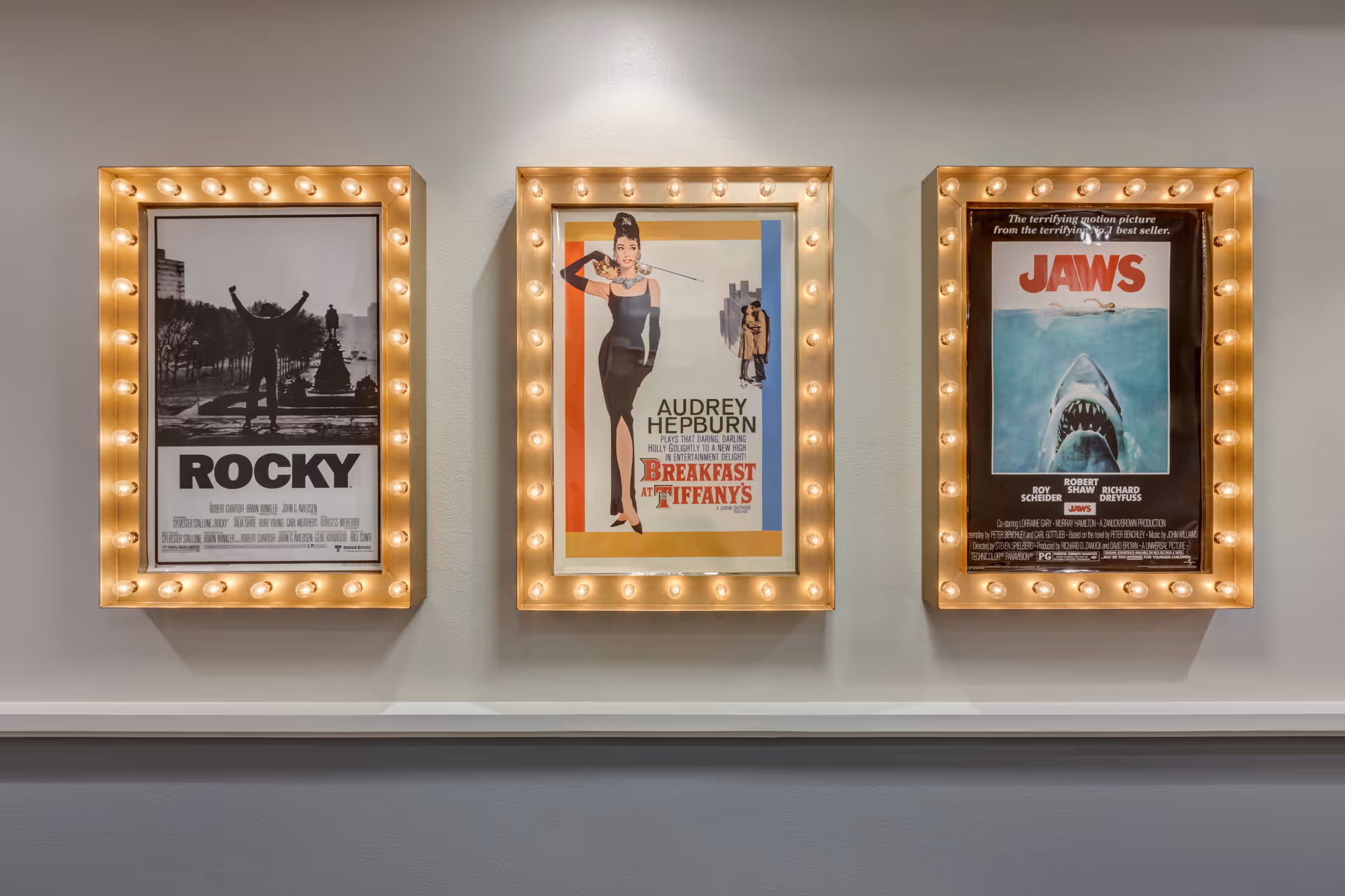 Three illuminated movie posters mounted on a wall. From left to right, the posters are for the films Rocky, Breakfast at Tiffany's featuring Audrey Hepburn, and Jaws.