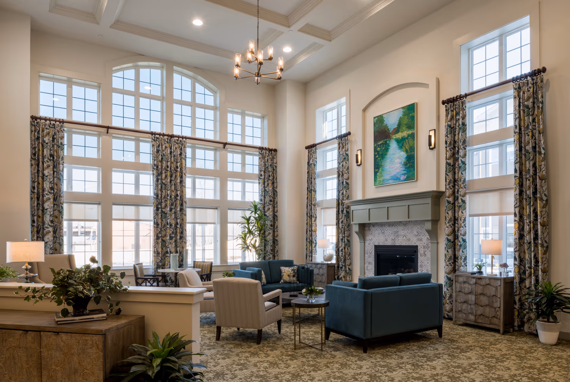 A spacious and well-lit living room area in a senior living facility featuring large windows with floral curtains, a fireplace with a painting above it, comfortable seating including blue sofas and beige armchairs, side tables with lamps, and decorative plants.