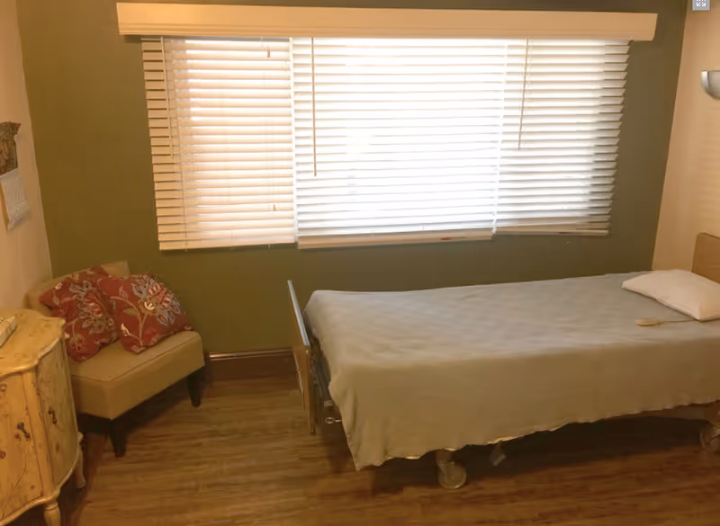 A simple bedroom with a single bed covered with a beige blanket, a pillow, and a remote control on the bed. There is a small beige armchair with two red patterned cushions next to a small wooden cabinet. A large window with white blinds covers most of the green wall behind the bed.