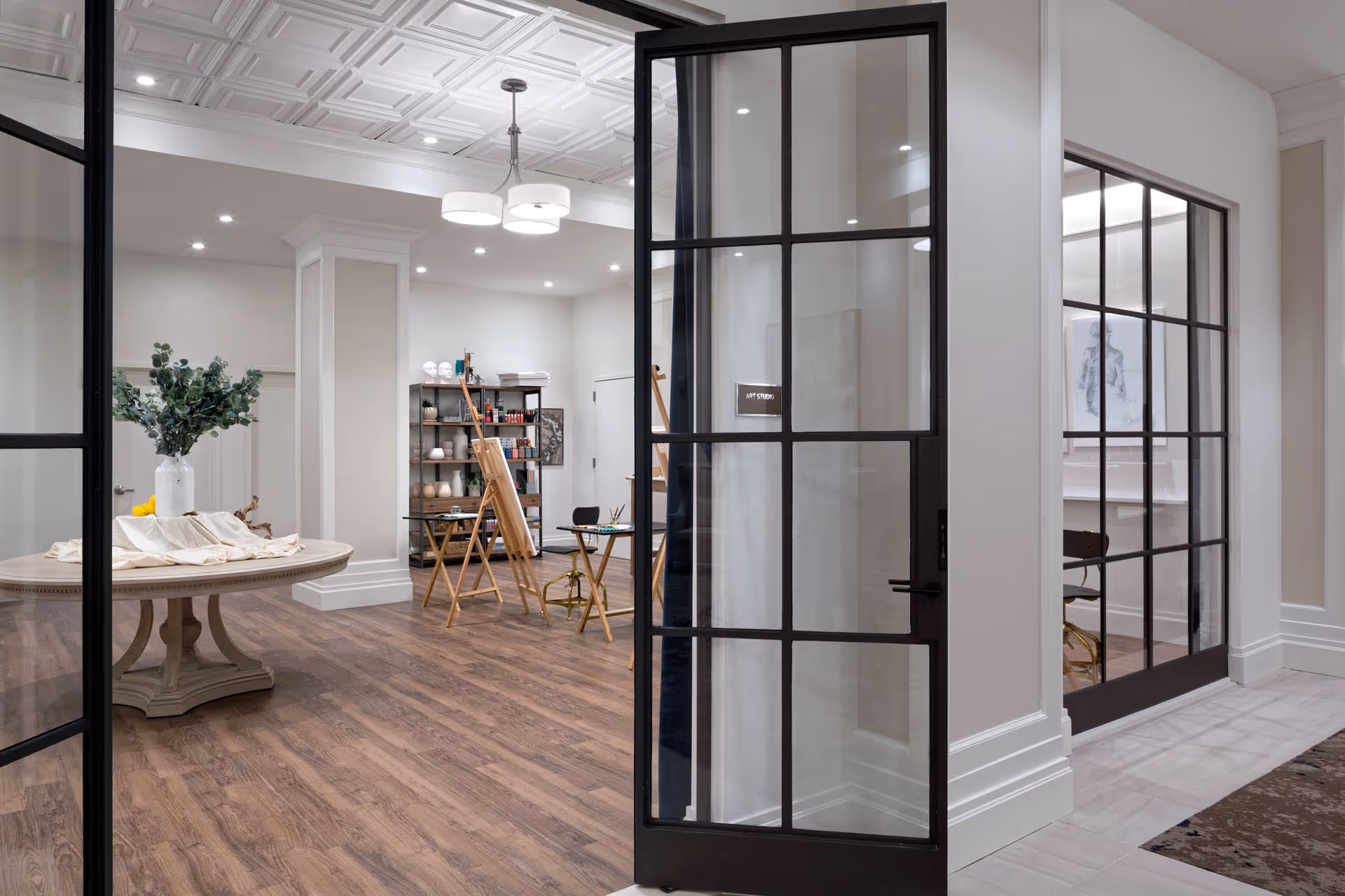 Interior view of an art studio room with wooden flooring, a round table with a vase of greenery, easels, shelves with art supplies, and large glass doors with black frames.