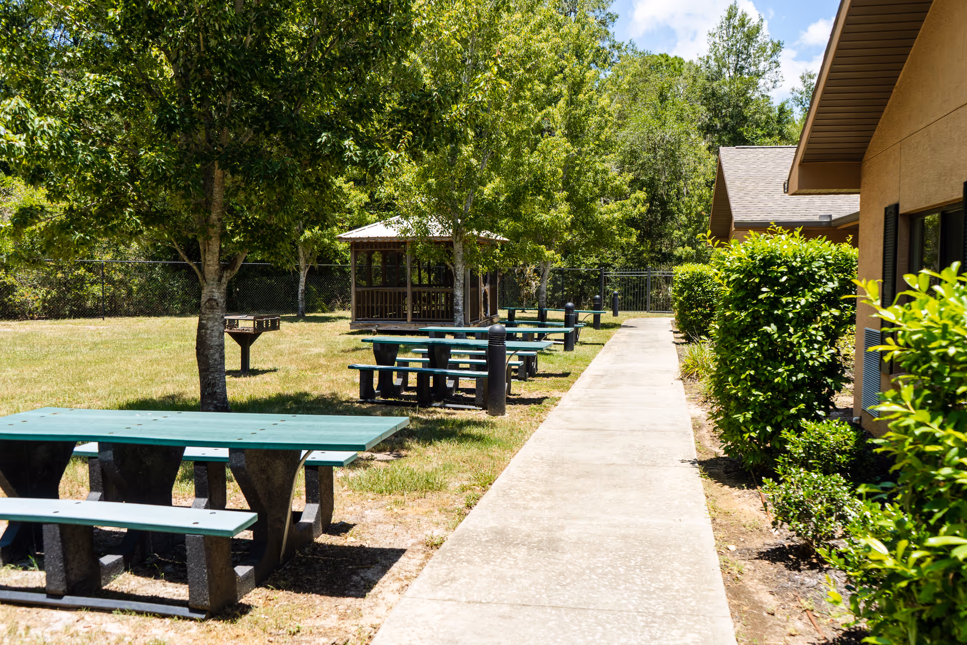 Outdoor area with green picnic tables and benches along a concrete pathway. There are trees providing shade, a small gazebo in the background, and bushes lining the side of a building under a partly cloudy sky.