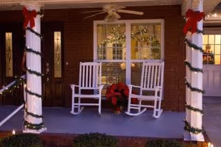 Front porch with two white rocking chairs, garland-wrapped columns with red bows, and a poinsettia between the chairs.