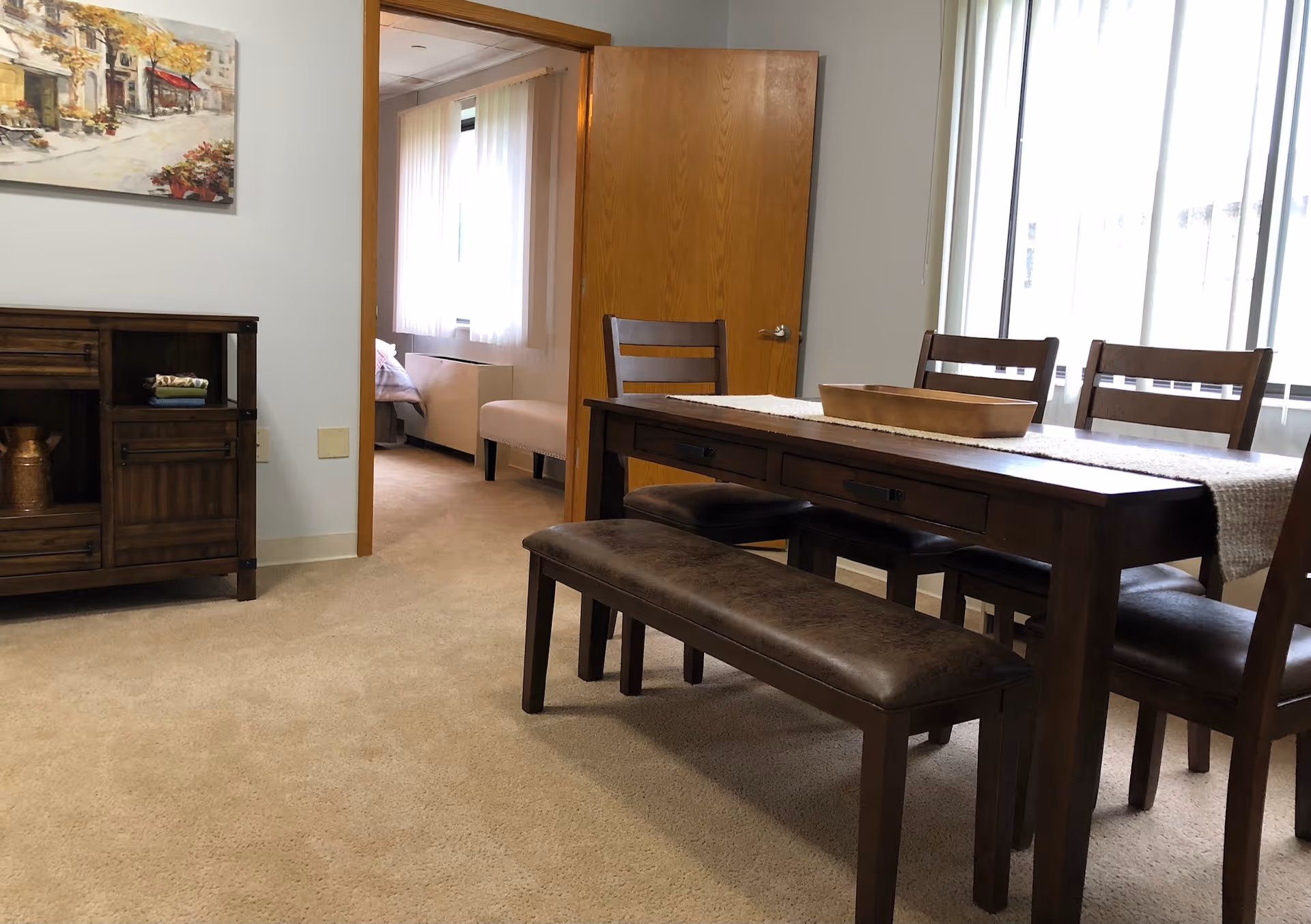 A dining room with a wooden table, four chairs, and a bench. The table has a white runner and a wooden tray on top. To the left, there is a wooden cabinet with decorative items and a painting above it. An open door reveals a bedroom with a bed and a window with sheer curtains. The room is carpeted and well-lit by natural light from large windows.