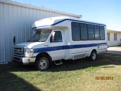 Ness County Hospital shuttle bus with blue stripe parked beside a white building.