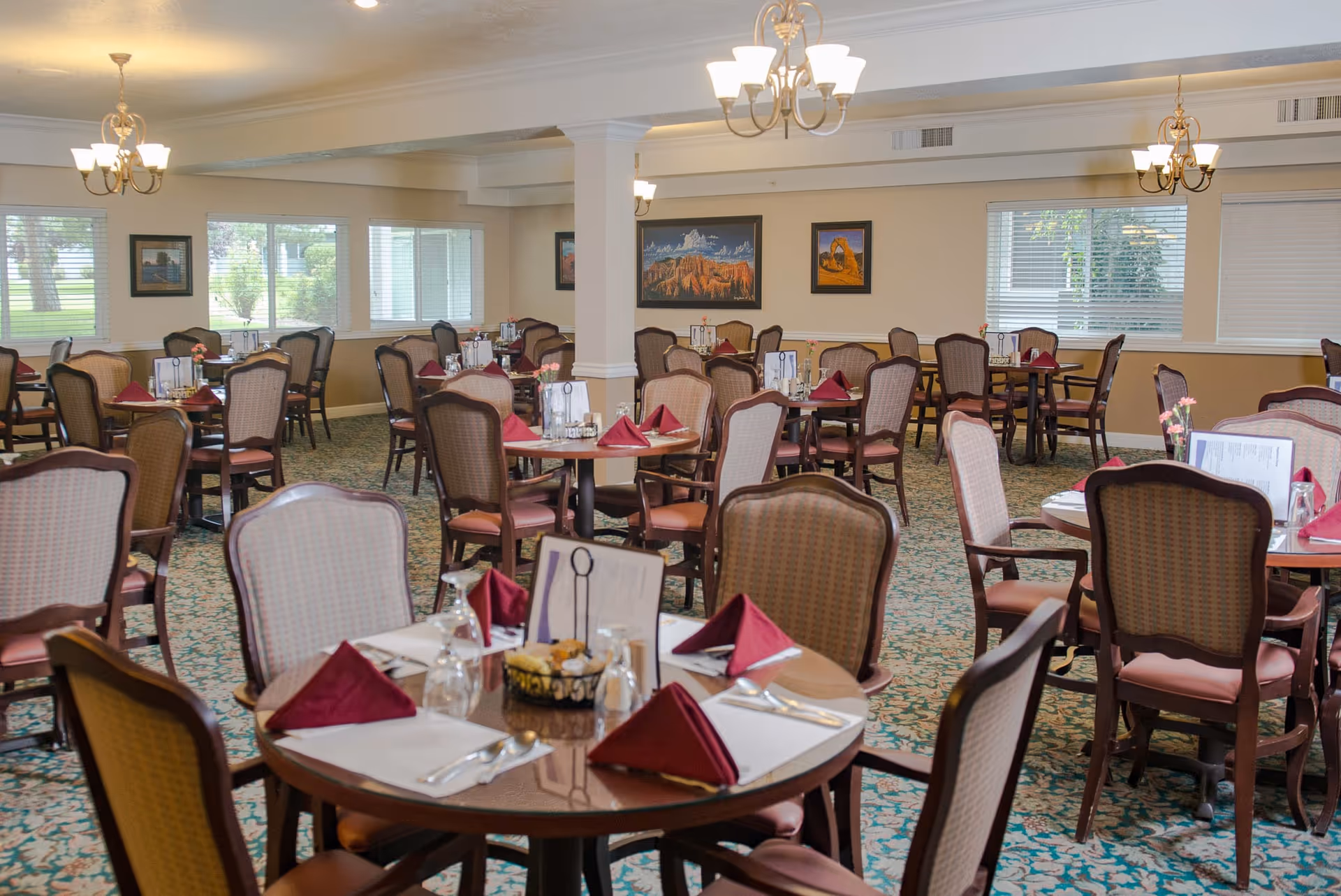 A spacious dining room with multiple round tables set with burgundy napkins, menus, glassware, and silverware. The room has patterned carpet, beige walls with framed landscape paintings, large windows with blinds, and chandeliers hanging from the ceiling.