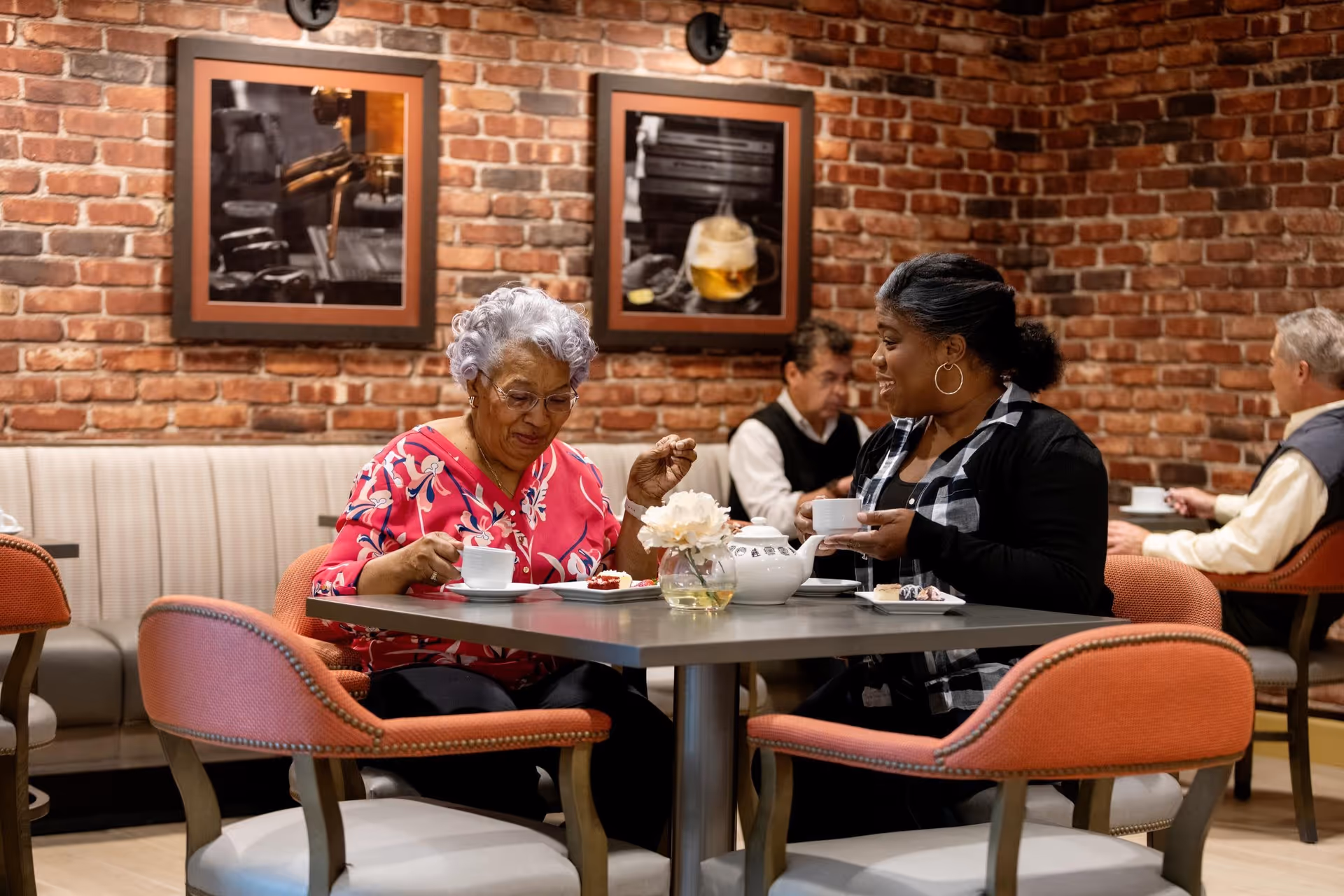 Two women sitting at a table in a cozy dining area with brick walls, enjoying tea and desserts. There are framed pictures on the wall behind them and two men seated at another table in the background.