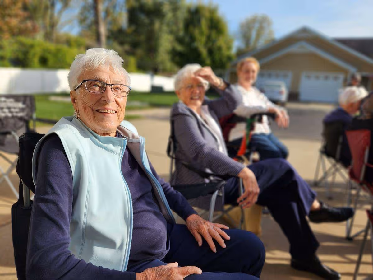 A group of elderly women sitting outdoors on folding chairs, enjoying a sunny day with trees and a house visible in the background.