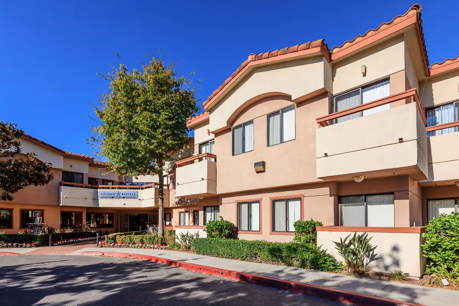 Exterior view of the Studio Royale senior living facility building with beige and light brown walls, red-tiled roof, balconies, windows, a tree, and landscaped bushes under a clear blue sky.