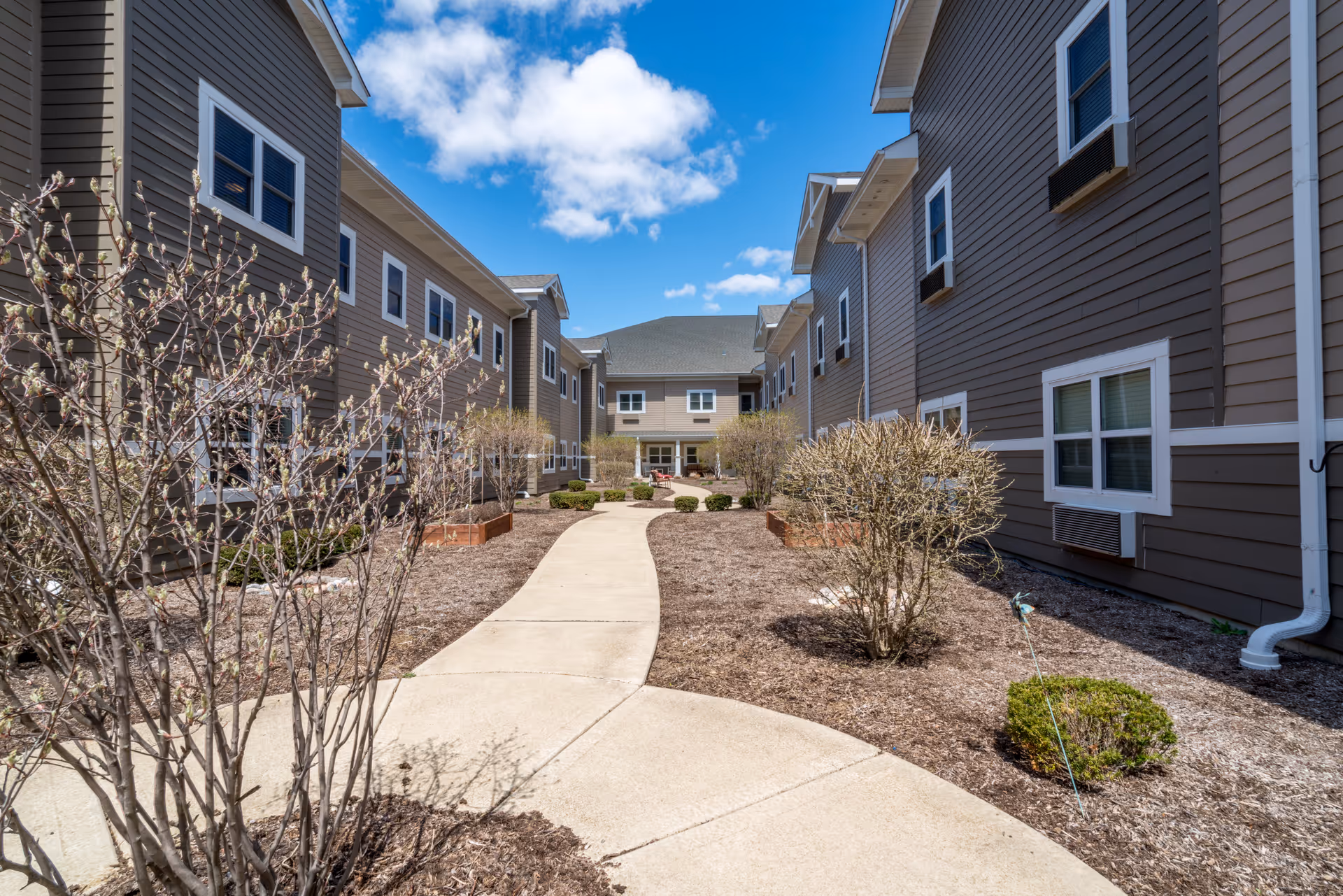 A paved walkway through a landscaped courtyard between two brown multi-story residential buildings under a blue sky.