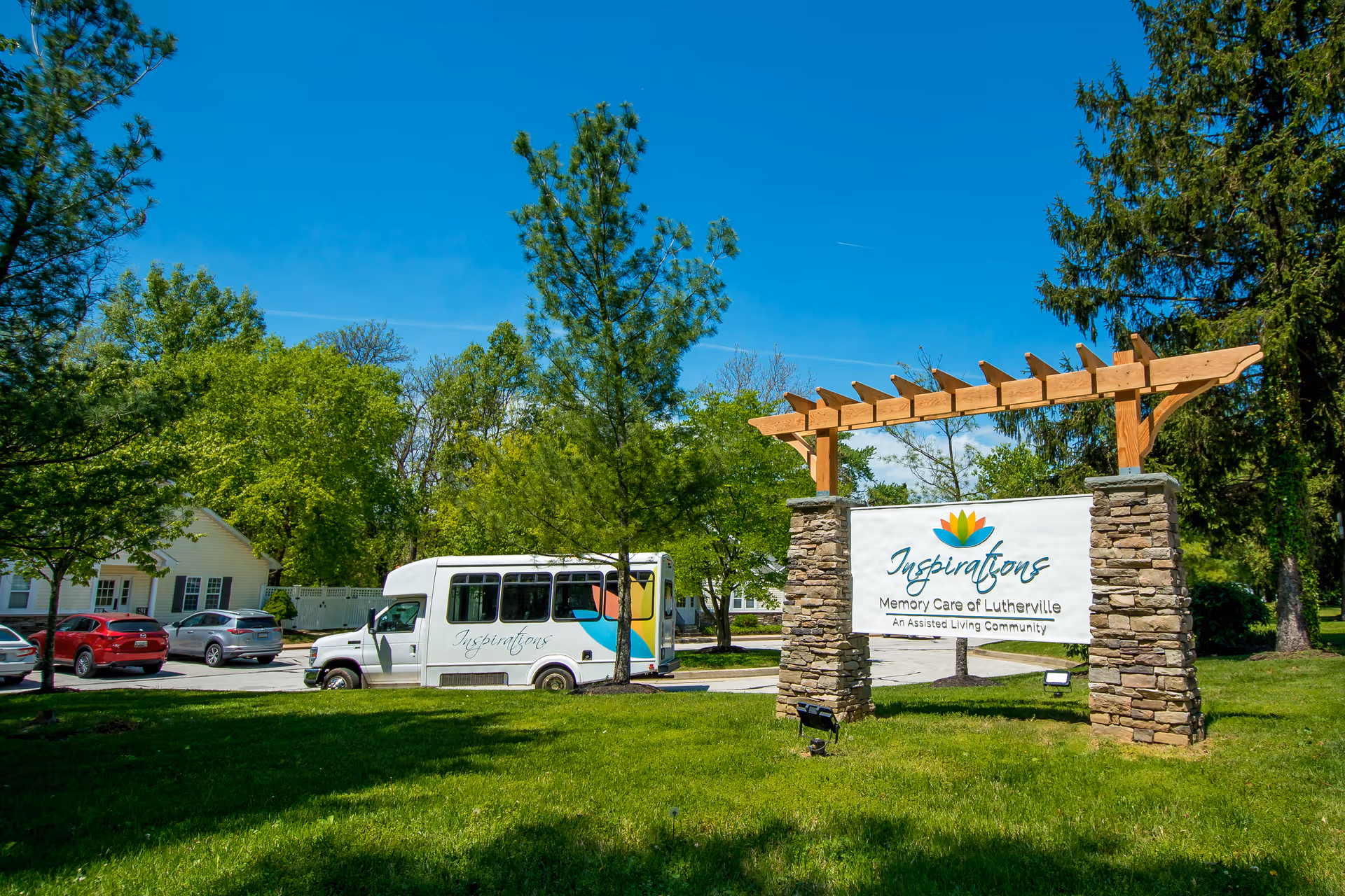 Outdoor view of Inspirations Memory Care of Lutherville featuring a large sign with the facility's name and logo supported by stone pillars and wooden beams. A white shuttle bus with the Inspirations logo is parked nearby, surrounded by green trees and a clear blue sky.