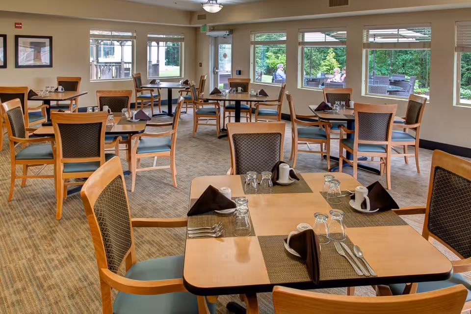 Dining room with neatly set wooden tables and chairs and large windows showing an outdoor patio.