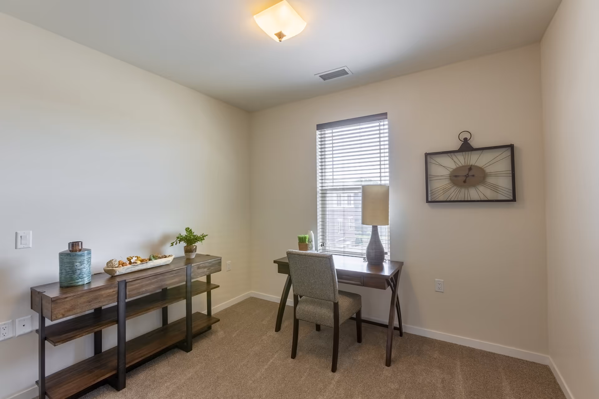A small, simple room with beige walls and carpeted floor featuring a wooden desk with a chair in front of a window with blinds. On the desk is a lamp and some small decorative items. To the left is a wooden console table with decorative items including a vase and a plant. A large wall clock is mounted on the wall to the right of the window.