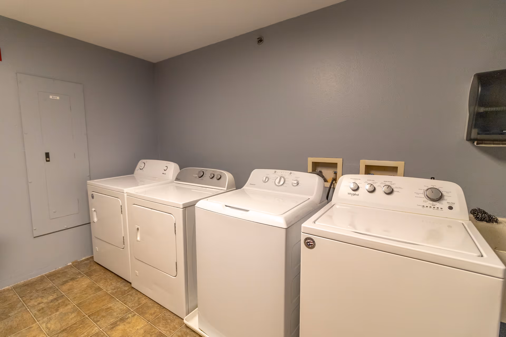 Laundry room with two white washing machines and two white dryers lined up against a gray wall, with a tiled floor and a paper towel dispenser mounted on the wall.