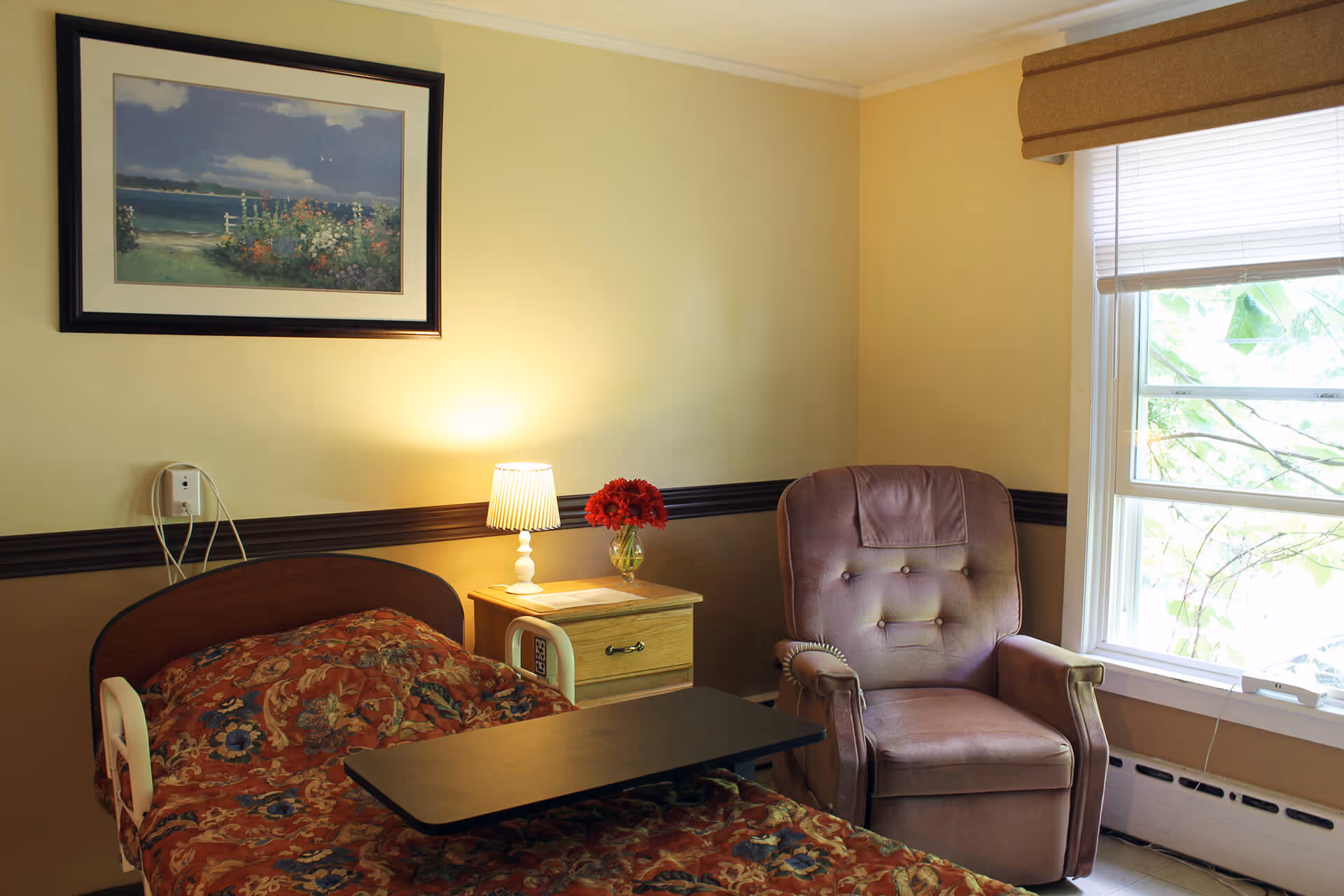 A cozy room with a single bed covered in a floral patterned blanket, a wooden nightstand with a lit lamp and a vase of red flowers, a comfortable upholstered armchair, and a window with blinds partially open letting in natural light. A framed painting of a seaside landscape hangs on the wall above the bed.