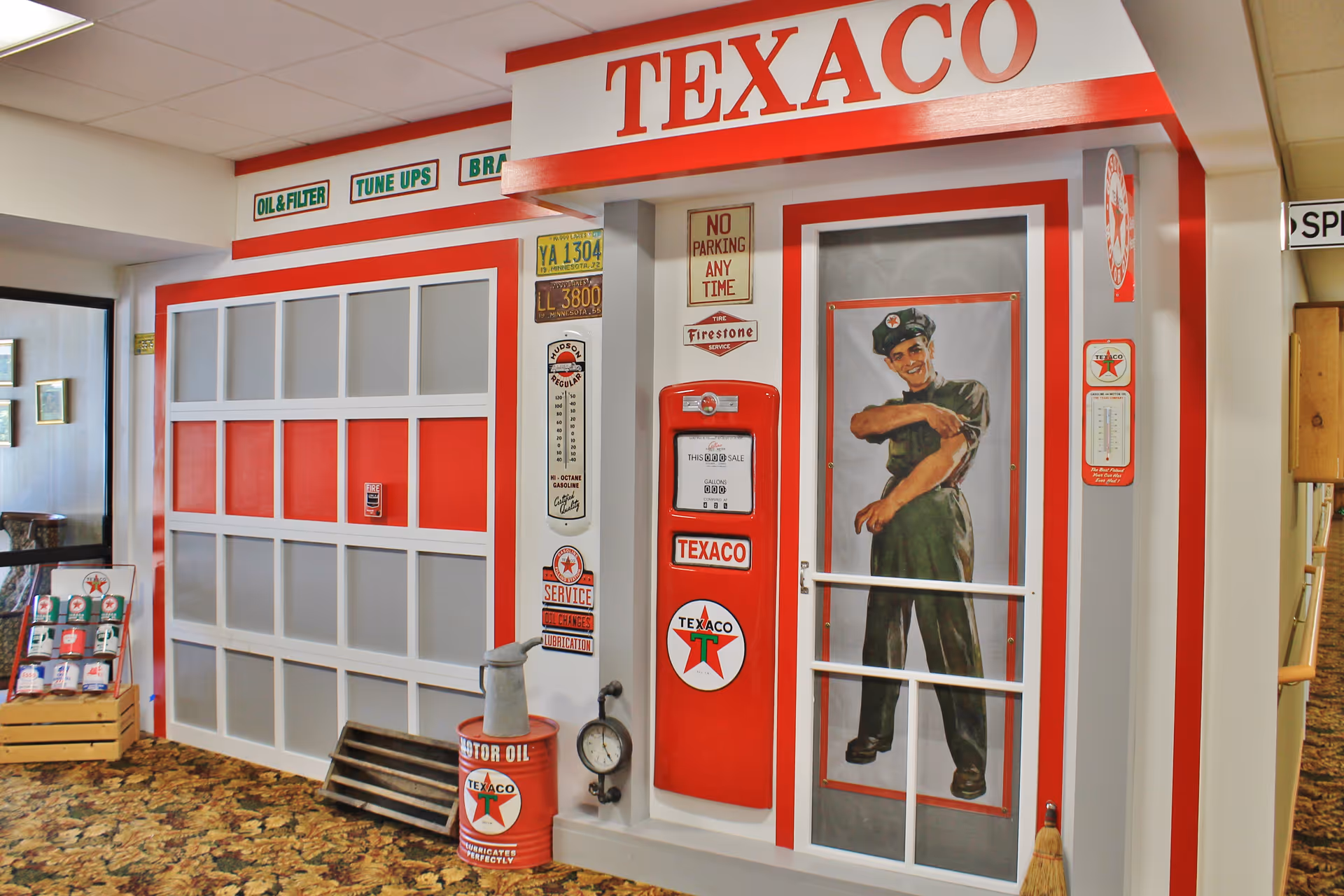 Interior view of a room decorated to resemble a vintage Texaco gas station, featuring a red and white garage door, a Texaco gas pump, various Texaco signs, a barrel labeled motor oil, and a large poster of a smiling gas station attendant rolling up his sleeve.