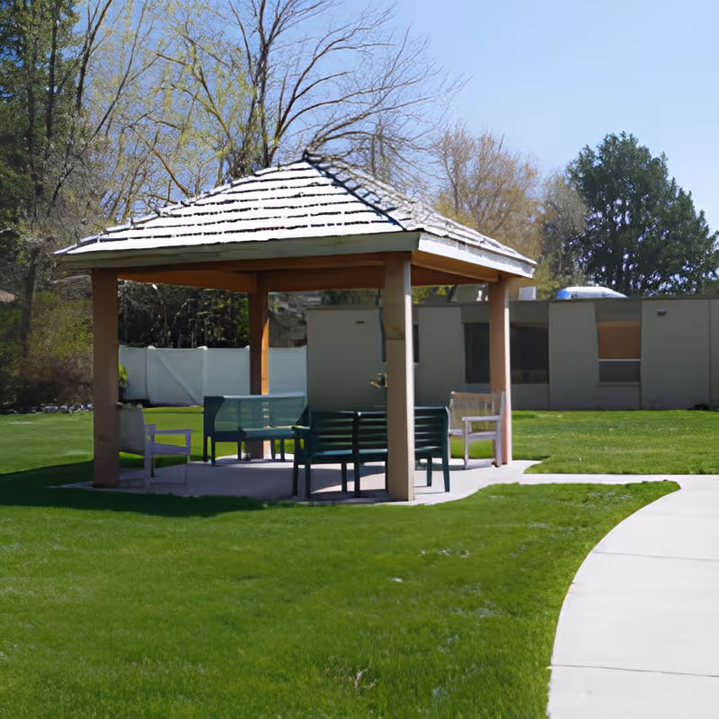 A wooden gazebo with benches on a grassy lawn beside a paved walkway, with a low building and trees in the background.