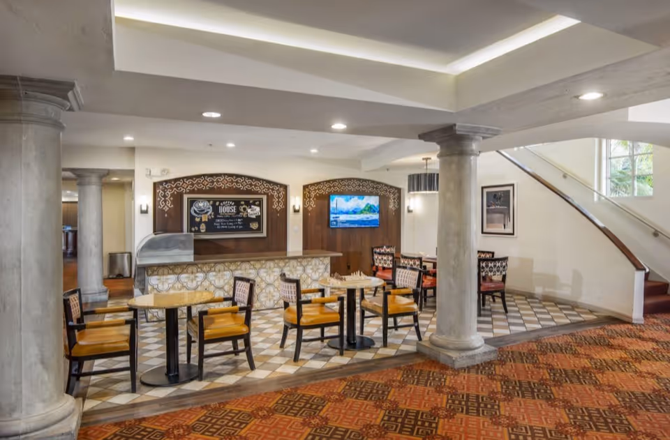 Interior view of a seating area in a senior living facility with several tables and chairs arranged on a tiled floor. There are two large columns, a counter with decorative tiles, a wall-mounted TV, and a staircase with a window letting in natural light.