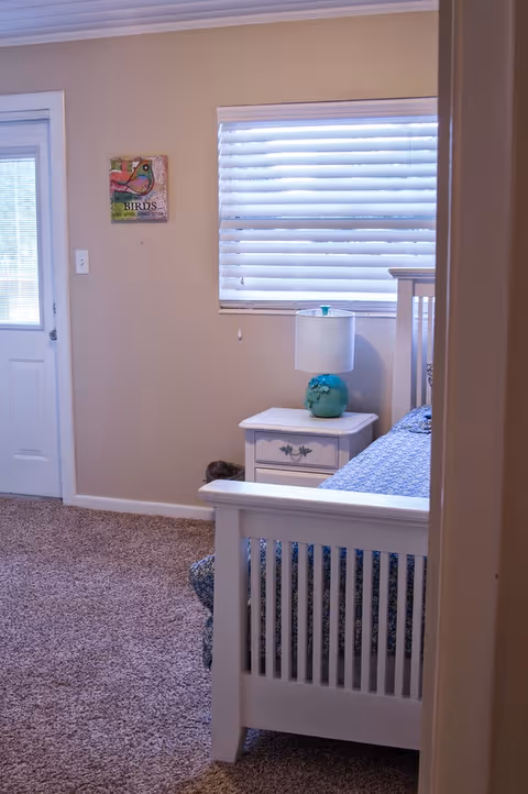 A cozy bedroom with beige walls and carpeted floor. There is a white wooden bed with a blue patterned bedspread partially visible. Next to the bed is a white nightstand with a turquoise lamp. A window with white blinds is above the nightstand, and a door with a window is on the left wall. A small colorful bird-themed wall decoration is hung near the door.