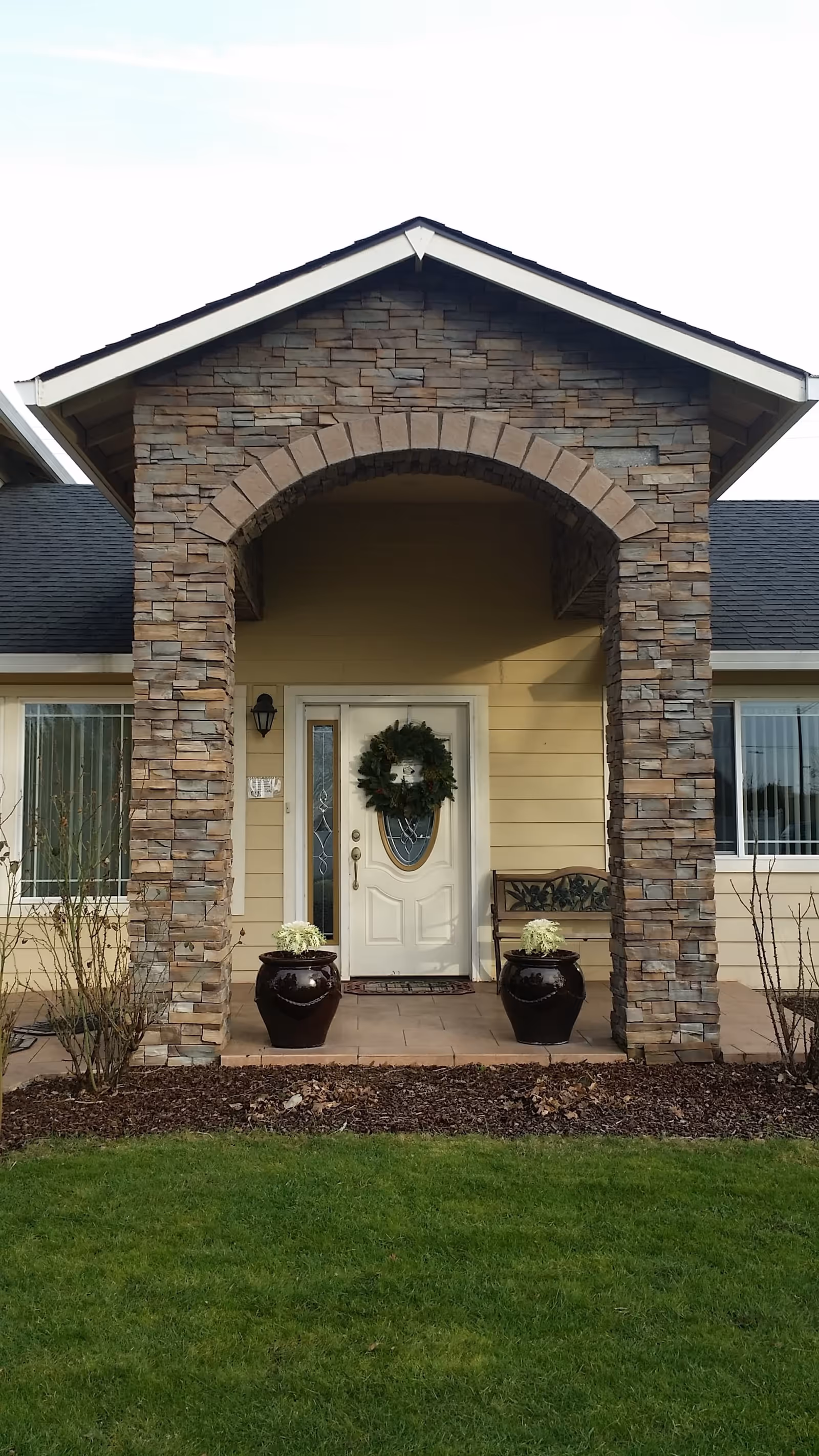 Front entrance of a building with a stone archway, a white door decorated with a green wreath, two large dark planters with white flowers on either side of the door, a bench to the right, and windows with vertical blinds on both sides.