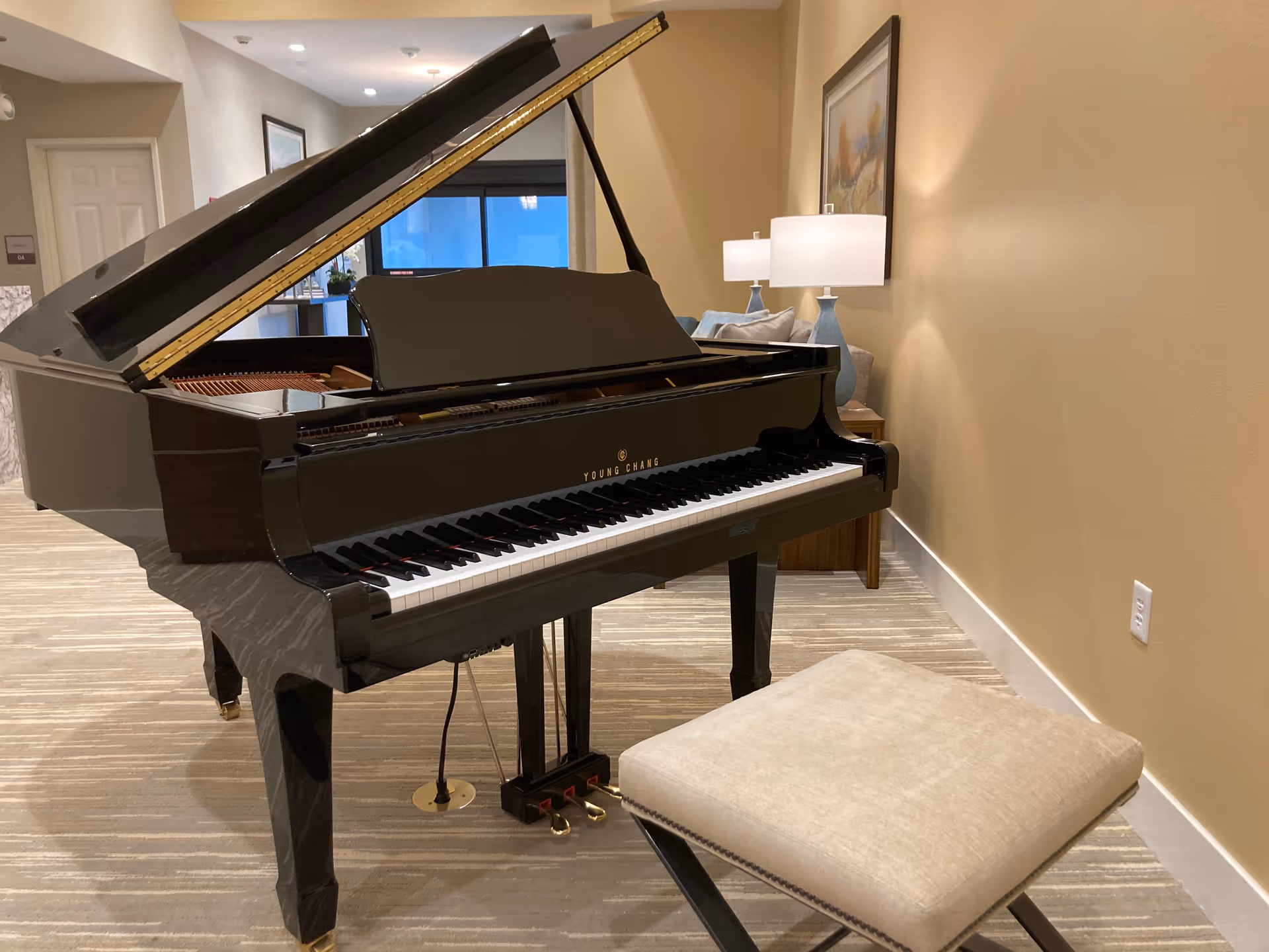 A black grand piano with its lid open is placed in a carpeted room with beige walls. In front of the piano is a cushioned beige piano bench. Behind the piano, there is a wooden side table with two blue lamps and a framed painting on the wall. A window with a dark frame is visible in the background.