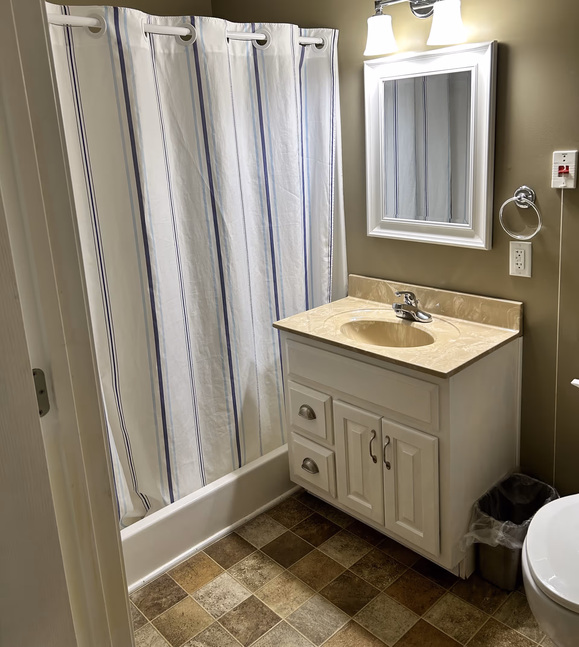 A bathroom with a white vanity cabinet topped with a beige marble countertop and an oval sink. Above the sink is a white-framed mirror with a light fixture above it. To the left is a bathtub with a white shower curtain featuring vertical blue stripes. The floor has a multi-colored tile pattern, and a small trash can is visible next to the toilet on the right.