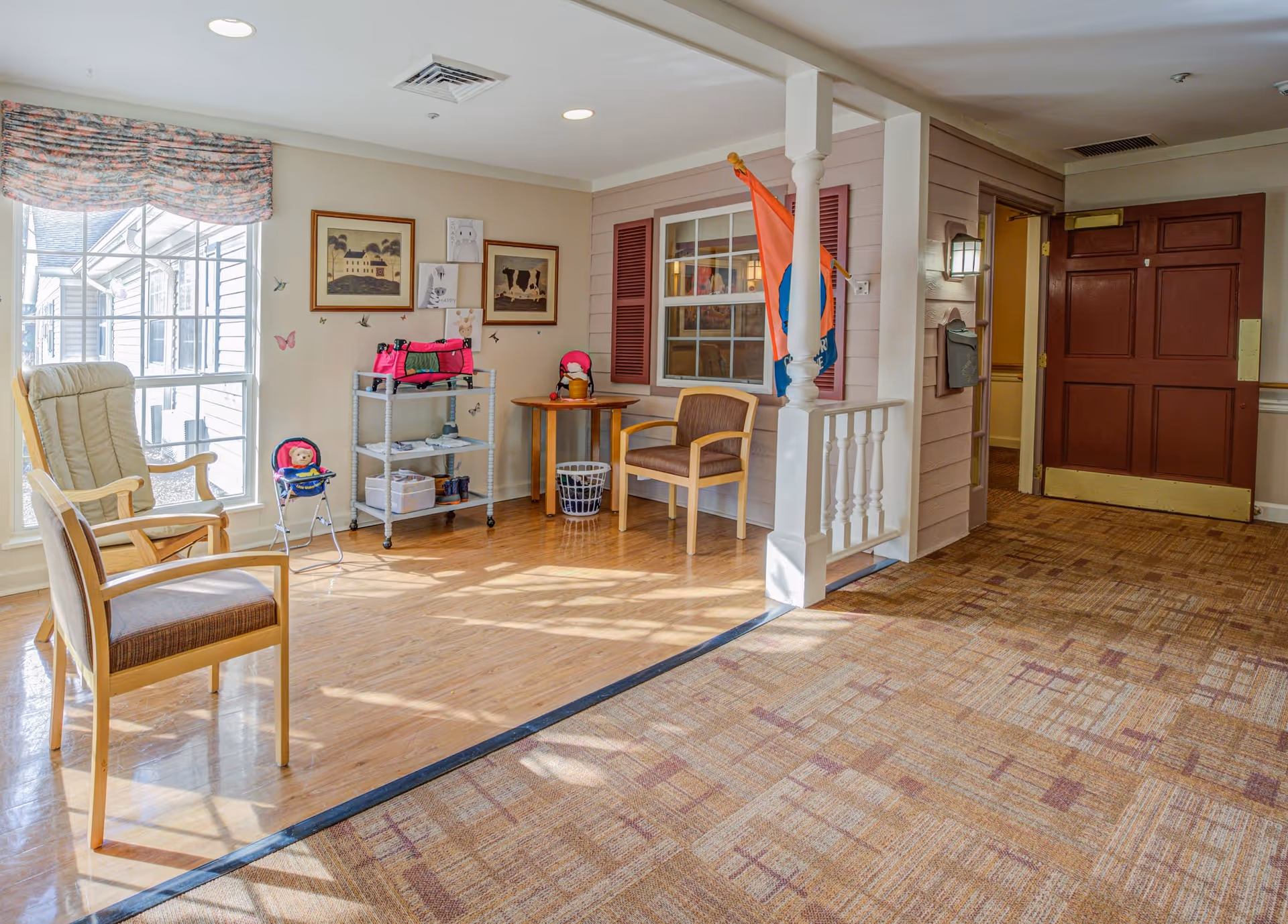 A bright and cozy sitting area in a senior living facility with two wooden chairs, a rocking chair, a small table, and a metal cart with various items. The space has large windows with floral curtains, framed artwork on the walls, and a flag hanging near a window with red shutters. The floor transitions from wood to carpet, and there is a dark red door in the background.