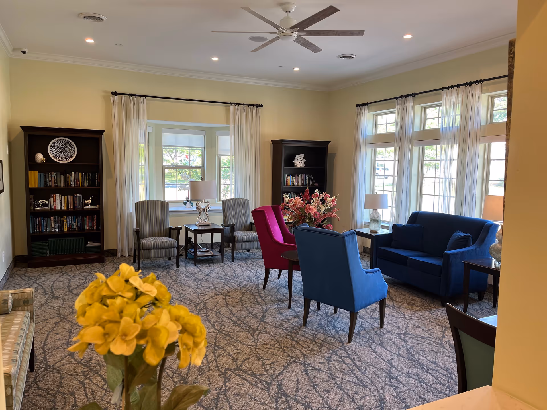 Bright common living room with armchairs, a blue sofa, bookshelves, and floral centerpieces by large windows.