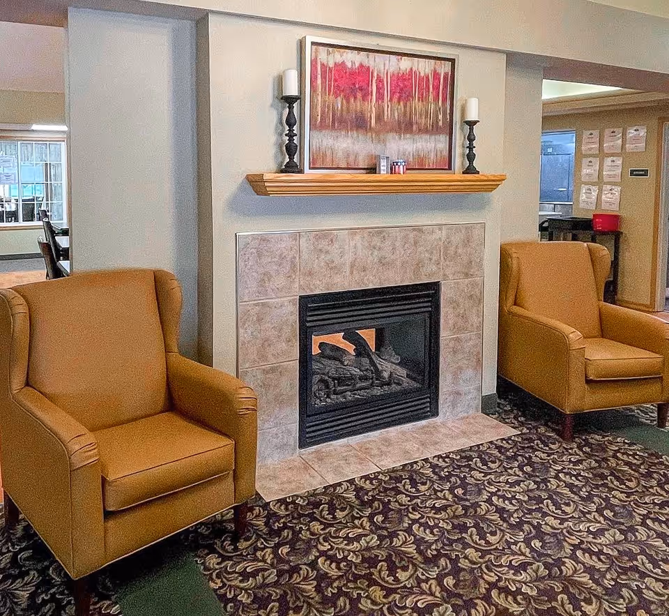 A cozy seating area with two tan armchairs positioned on either side of a tiled fireplace. Above the fireplace is a wooden mantel holding two black candle holders with white candles and a colorful painting featuring red and pink trees. The floor is covered with a patterned carpet, and the background shows part of a dining area and hallway.