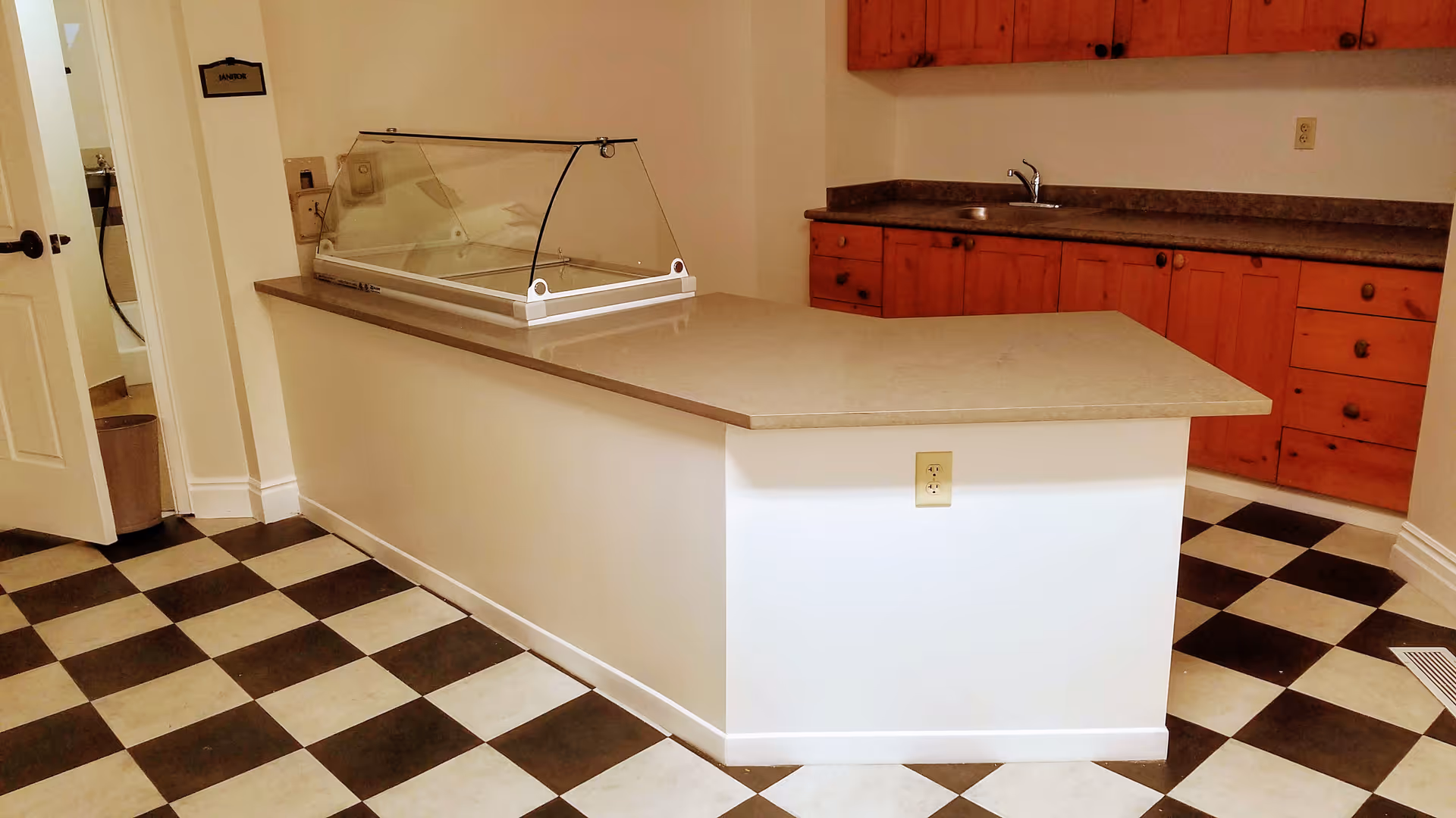 A serving counter with a glass sneeze guard in a kitchenette featuring checkered tile floor and wooden cabinets with a sink.