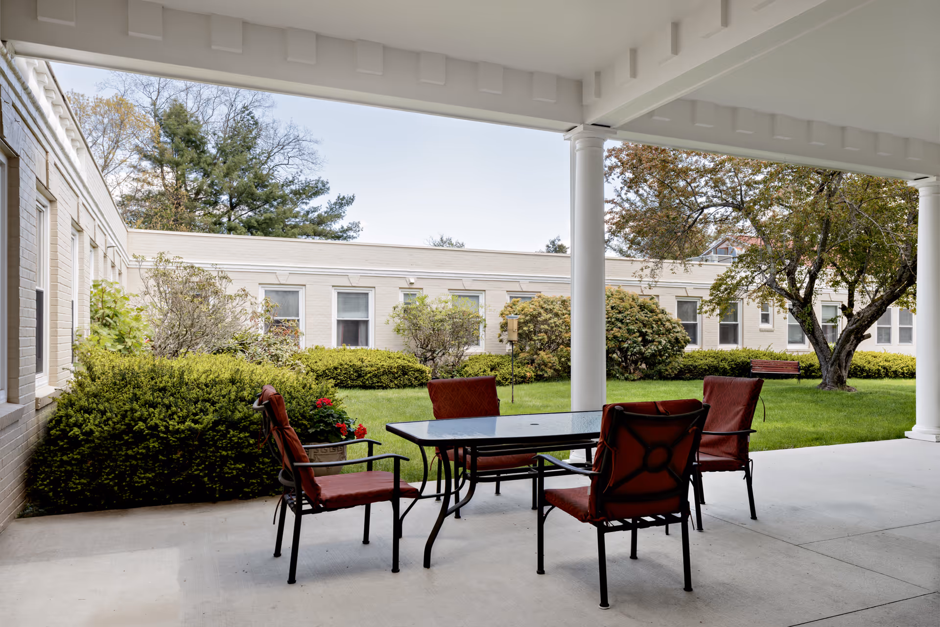 Covered outdoor patio area with a glass-top table and four red cushioned chairs. The patio overlooks a green lawn with bushes, trees, and a bench. The building with multiple windows surrounds the lawn area.