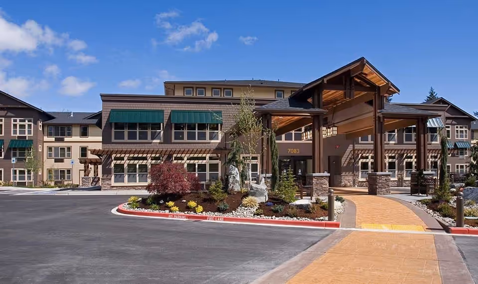 Exterior front entrance of a multi-story senior living building with a covered porte-cochère, landscaped island, and paved driveway.
