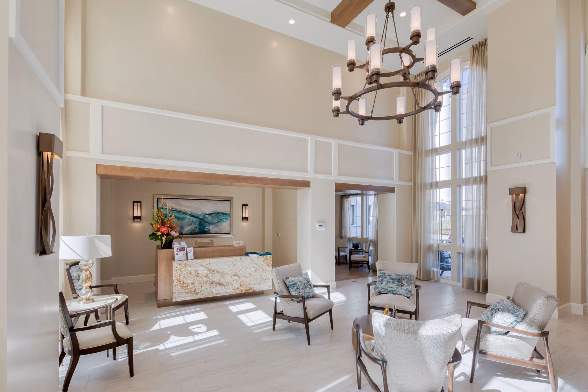 Bright and spacious senior living facility lobby with high ceilings, large windows with sheer curtains, a chandelier, a reception desk with a flower arrangement, and several cushioned chairs arranged in a seating area.