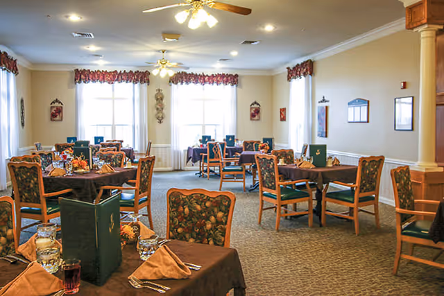 A dining room with multiple tables covered in brown tablecloths, each set with napkins, glasses, and menus. The room has large windows with floral valances, ceiling fans with lights, and beige walls with framed decorations.