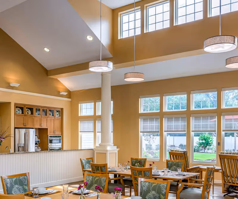 Bright dining area with multiple wooden tables and chairs featuring green floral cushions. The room has large windows letting in natural light, beige walls, and a high ceiling with hanging round light fixtures. A kitchen area with wooden cabinets and stainless steel appliances is visible in the background.