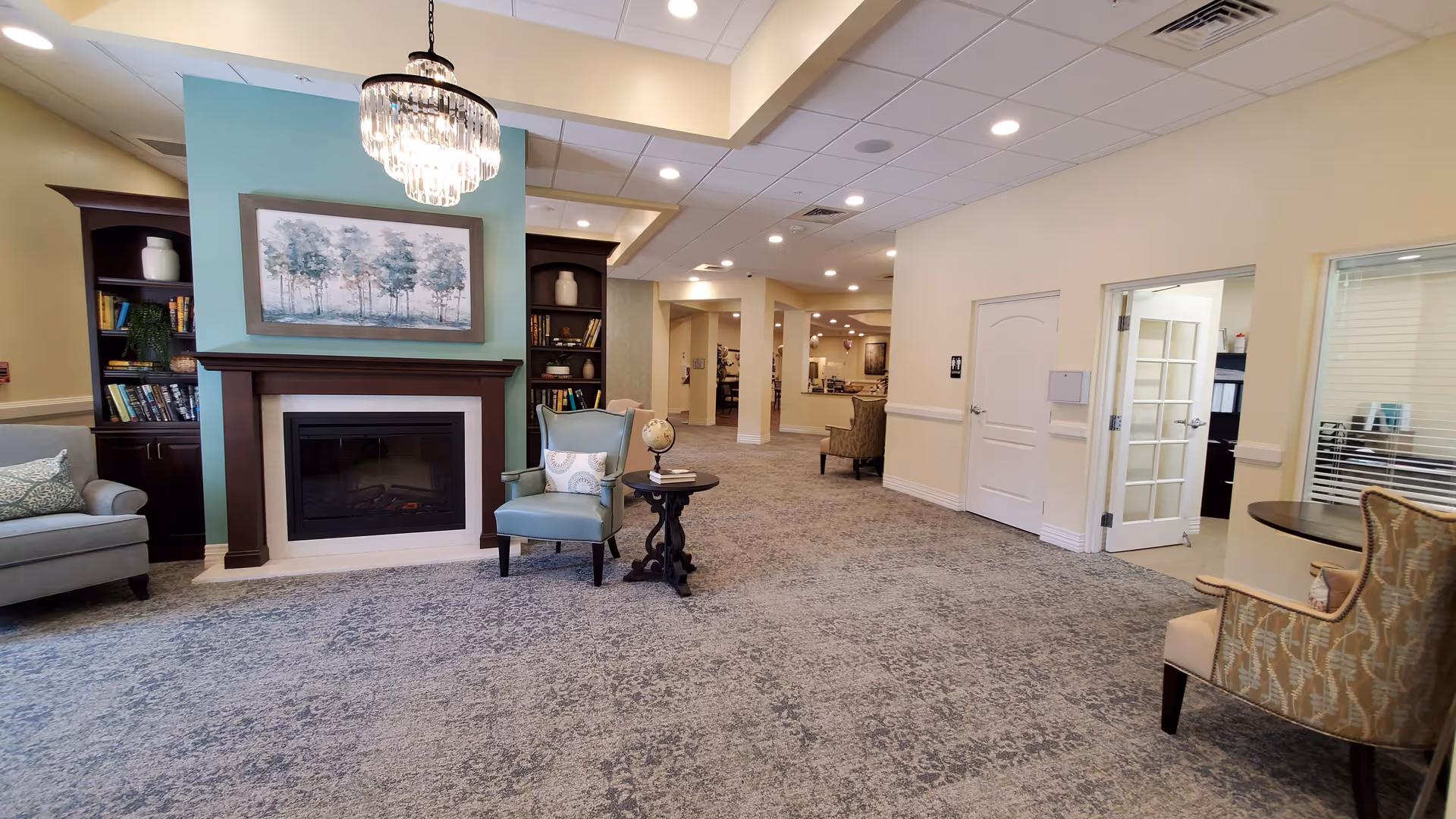 Spacious senior living common area with a fireplace, armchairs, built-in bookshelves, and a chandelier.