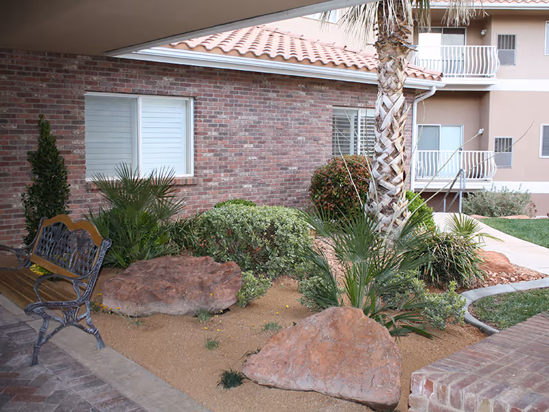 Outdoor garden area with desert landscaping including large rocks, various green plants, and a palm tree. There is a decorative metal and wood bench on the left side and a brick building with windows in the background. A walkway and part of a multi-story building with balconies are visible on the right.