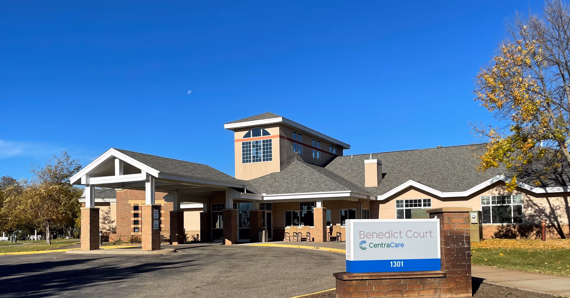 Front entrance of Benedict Court senior living building with a covered porte-cochère and sign under a clear blue sky.