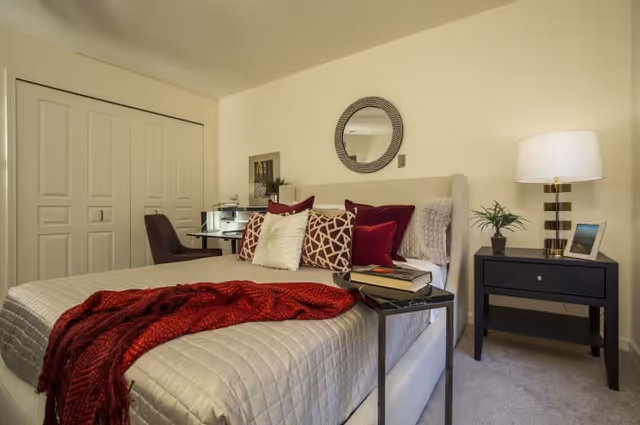 A cozy bedroom with a beige upholstered bed featuring a quilted bedspread, decorative pillows in white, burgundy, and patterned designs, and a red throw blanket. There is a small black side table with a lamp, a small plant, and a framed photo. A round mirror hangs on the wall above the bed. In the background, there is a desk with a chair and a closet with double doors.