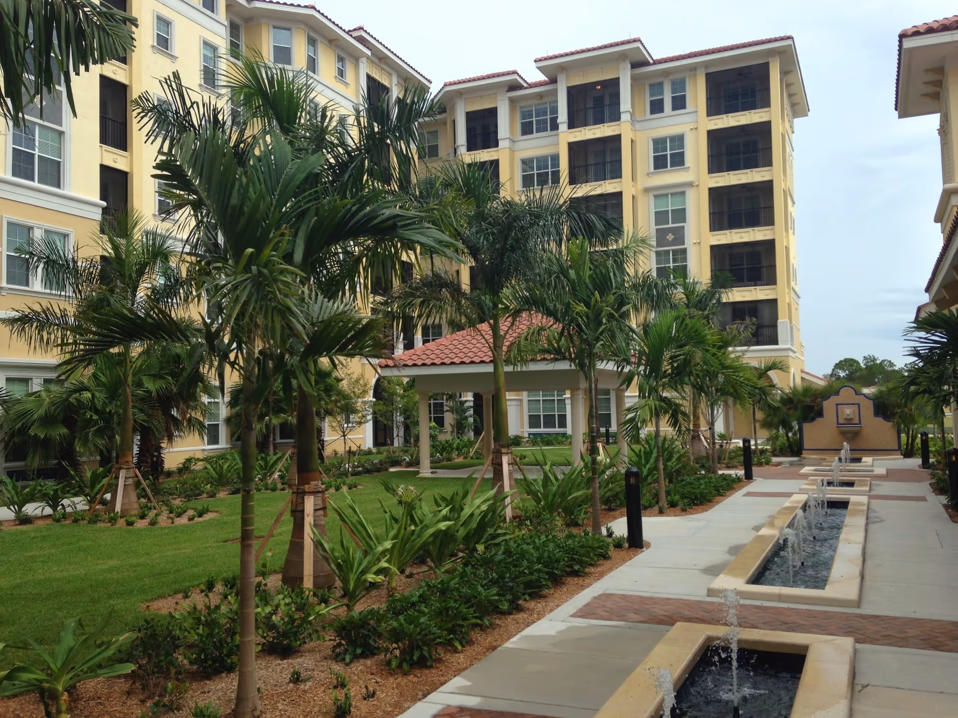 Outdoor courtyard area of a senior living facility with palm trees, landscaped garden beds, a walkway, and a series of rectangular water fountains. The multi-story building with yellow walls and white trim surrounds the courtyard.