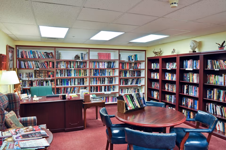 Cozy interior library/reading room with bookshelves lining the walls, a round table with chairs, and a desk with a lamp.