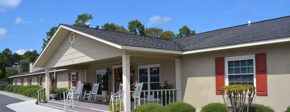 Exterior view of Southern Manor Senior Living building with beige walls, red window shutters, a covered porch with white rocking chairs, and neatly trimmed bushes under a clear blue sky.