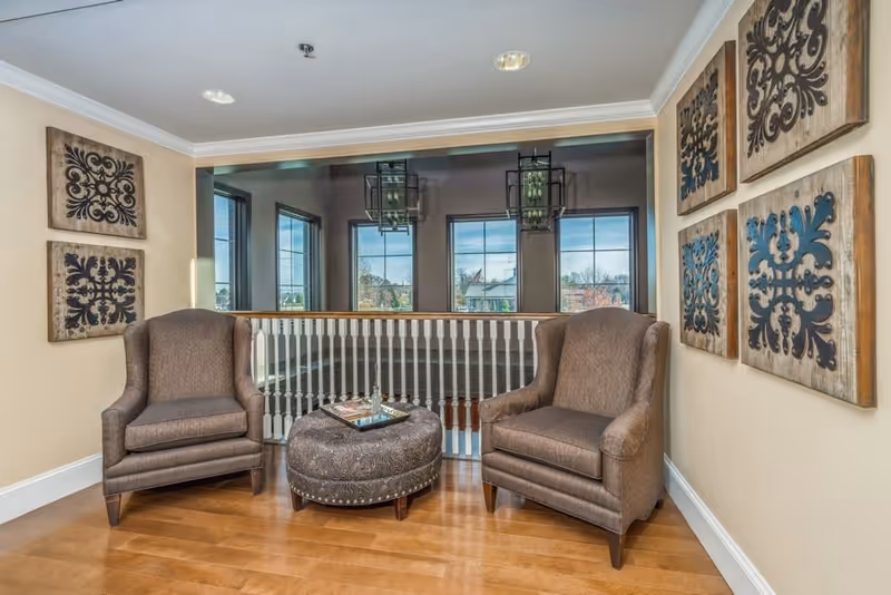 A cozy sitting area with two brown upholstered armchairs and a round ottoman with a tray on top, placed on a wooden floor. The walls are decorated with six square wooden art pieces featuring intricate black designs. Large windows in the background let in natural light, and a white railing separates the sitting area from the lower level.