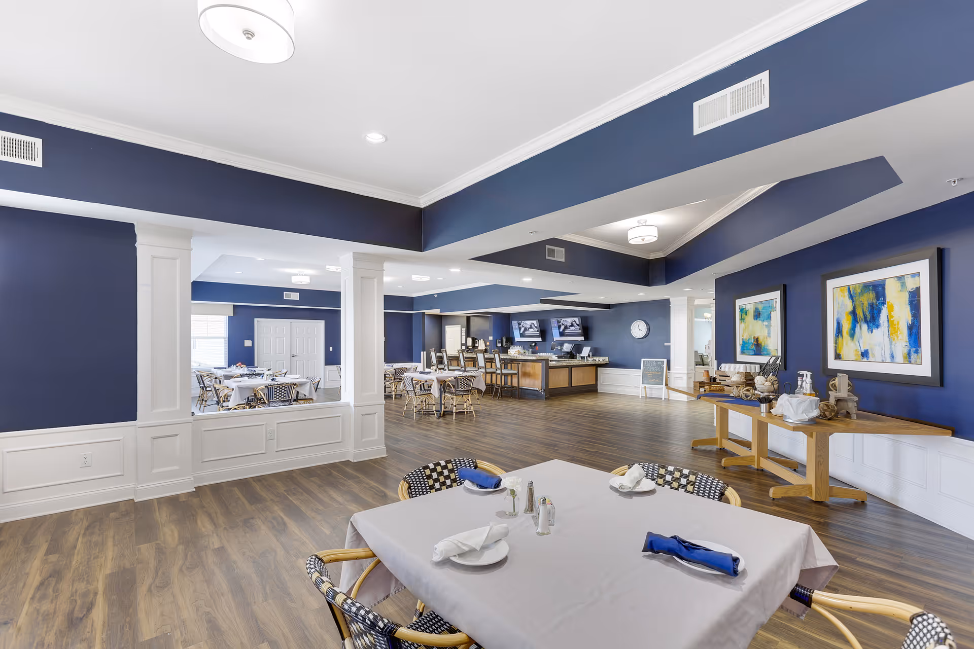 A spacious dining area with tables covered in white tablecloths, set with plates, napkins, and utensils. The room features dark blue walls with white trim, wooden flooring, and modern ceiling lights. There is a serving counter with two TV screens mounted above it and a side table with decorative items and artwork on the walls.