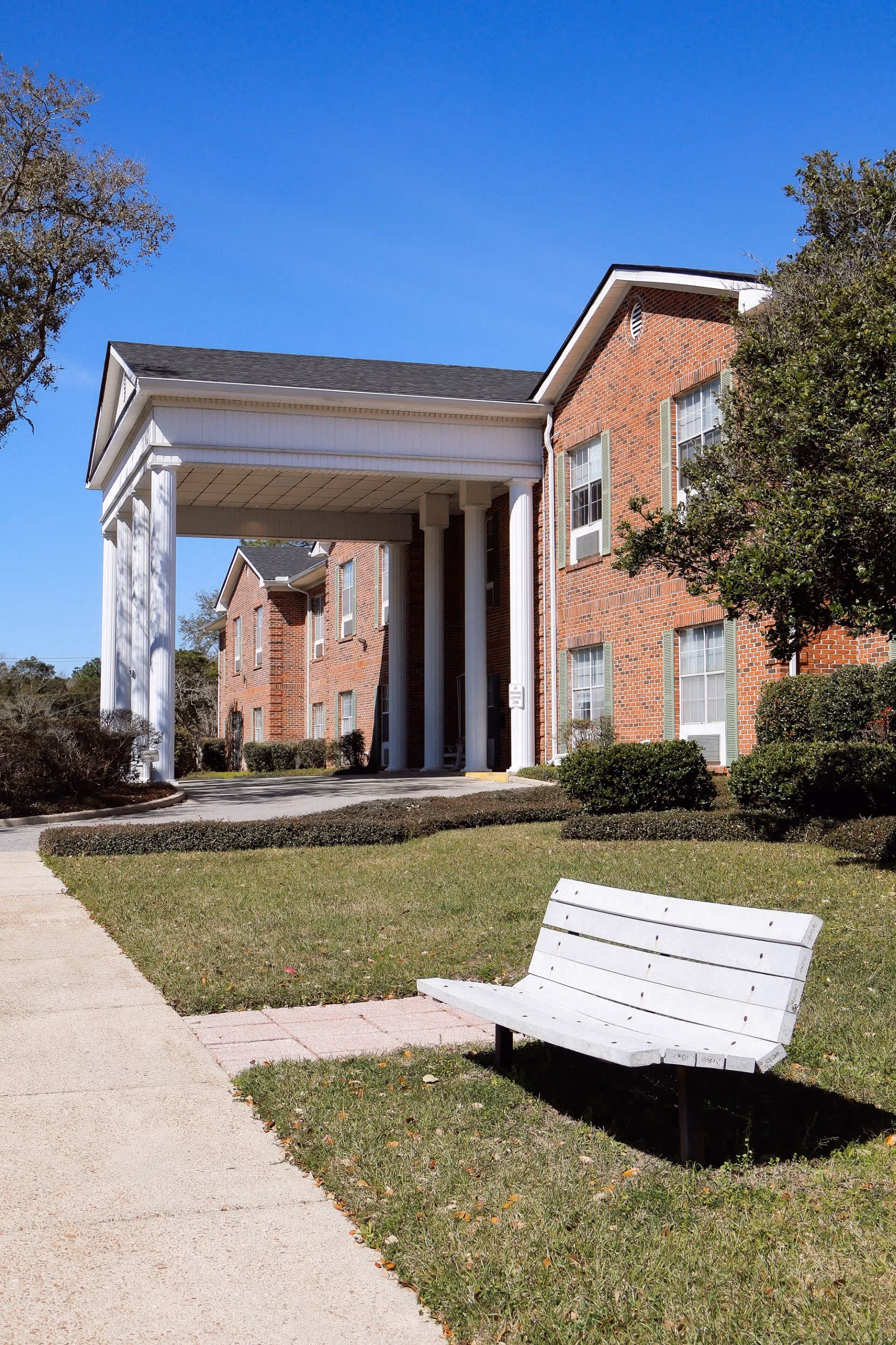 Exterior view of a senior living facility building with red brick walls, white columns supporting a covered entrance, green shrubs, a white bench on the grass, and a clear blue sky.