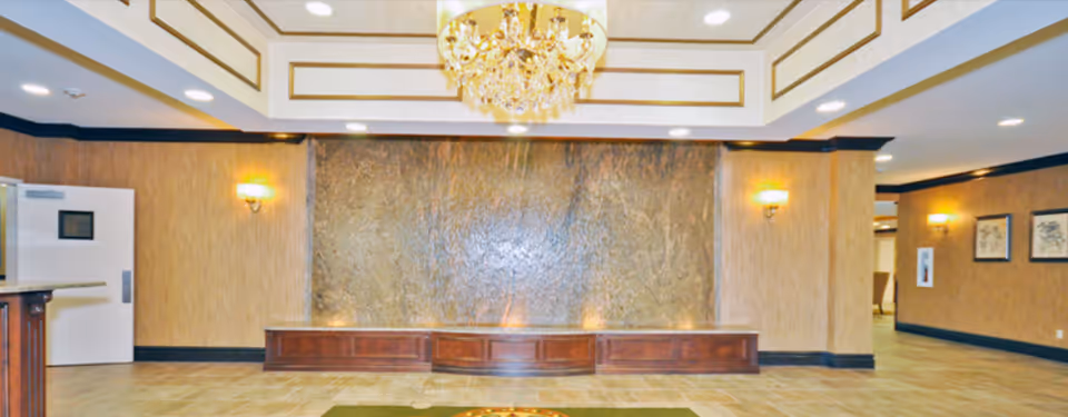 Interior view of a spacious room with a decorative water feature on the far wall, wooden paneling, wall sconces, a chandelier hanging from a recessed ceiling, and tiled flooring.