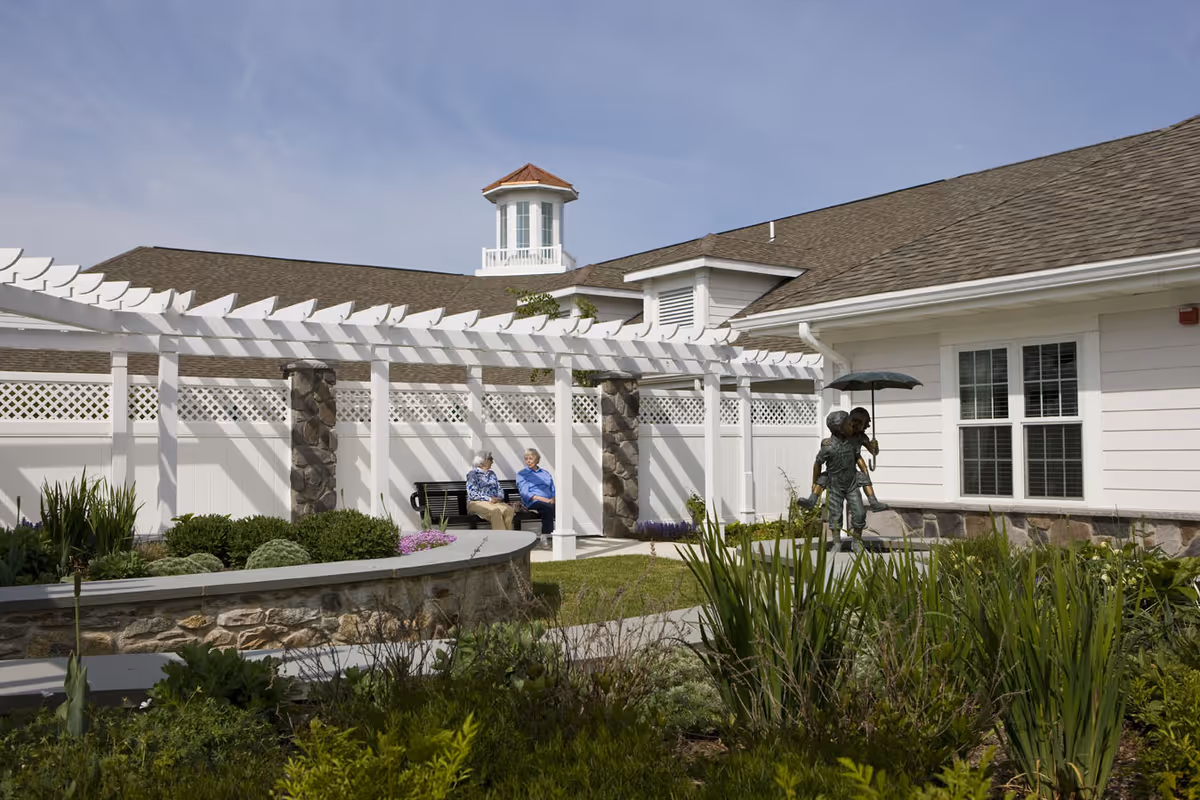 Outdoor garden area at StoneRidge Senior Living with two elderly people sitting on a bench under a white pergola, surrounded by greenery and flowers. A statue of two children holding an umbrella is visible near the building with white siding and a stone base.